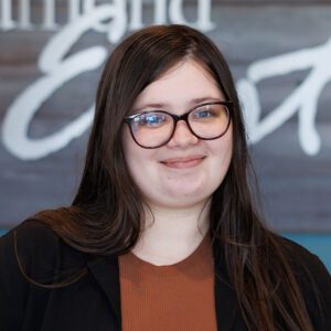 Jamie wearing a dark orange top and a black sweater smiling at the camera
