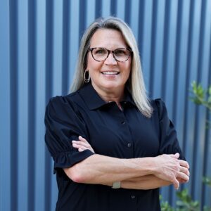 Kim G - female with shoulder length blond hair wearing a black blouse and dark glasses standing with arms crossed